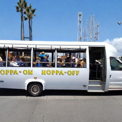 White open-sided tour bus with 'Hoppa-On Hoppa-Off' on the side, parked on a sunny street.