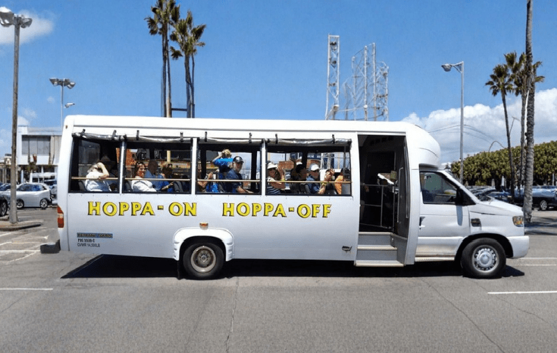 White open-sided tour bus with 'Hoppa-On Hoppa-Off' on the side, parked on a sunny street.