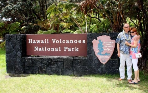 Couple standing by Hawaii Volcanoes National Park sign with foliage background.