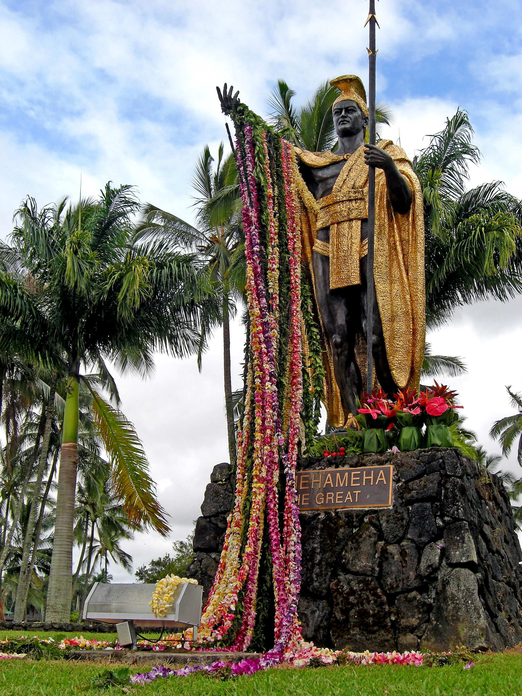 Statue of a standing figure draped with colorful leis, surrounded by palm trees.