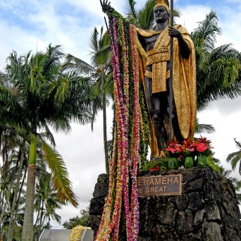 Statue of a standing figure draped with colorful leis, surrounded by palm trees.