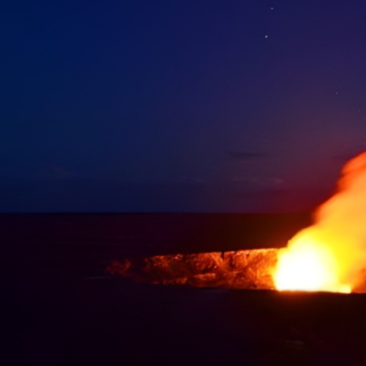 Lava glow in a crater against a dark sky with some trees visible.