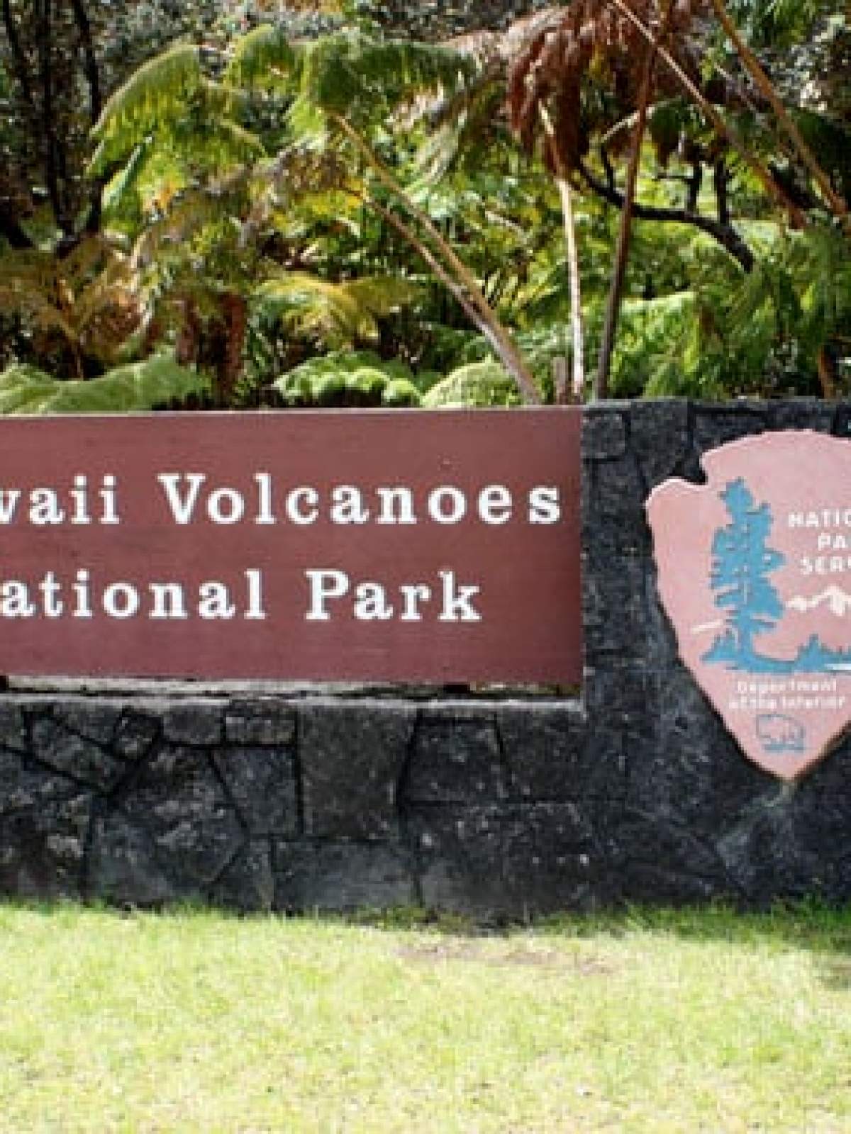 Couple stands by Hawaii Volcanoes National Park sign with lush foliage.
