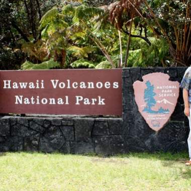 Couple stands by Hawaii Volcanoes National Park sign with lush foliage.