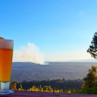 Tall glass of beer with orange slice on railing, overlooking a vast landscape and distant smoke plume.