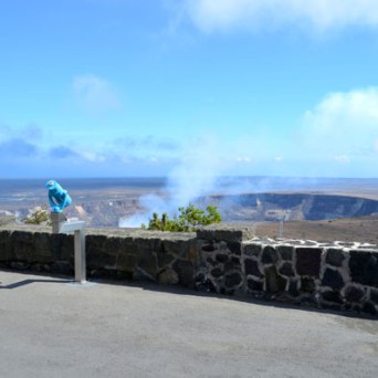 Stone wall view of distant smoking volcano crater under blue sky.