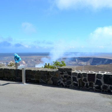 Stone wall view of distant smoking volcano crater under blue sky.