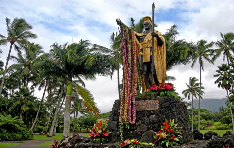Statue adorned with leis in tropical garden with palm trees and mountains in background.